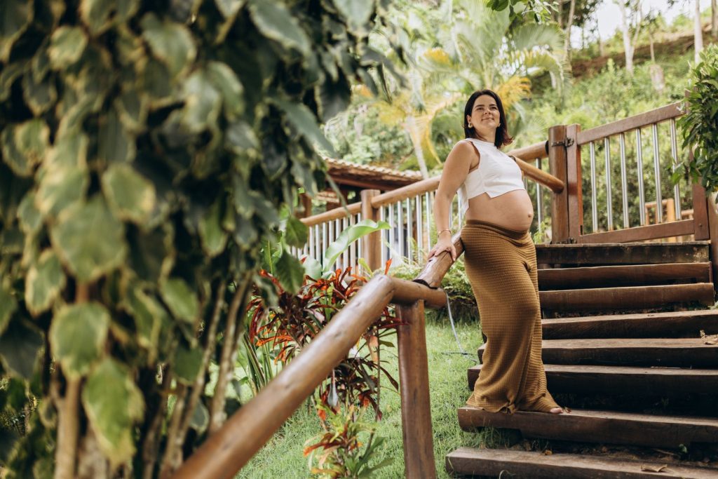 Pregnant woman in white top and brown skirt on stairs in lush greenery, enjoying a calm moment.