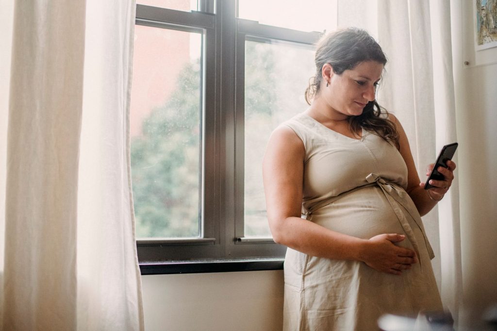 Adult pregnant lady checking information on phone while standing near window in room in daylight
