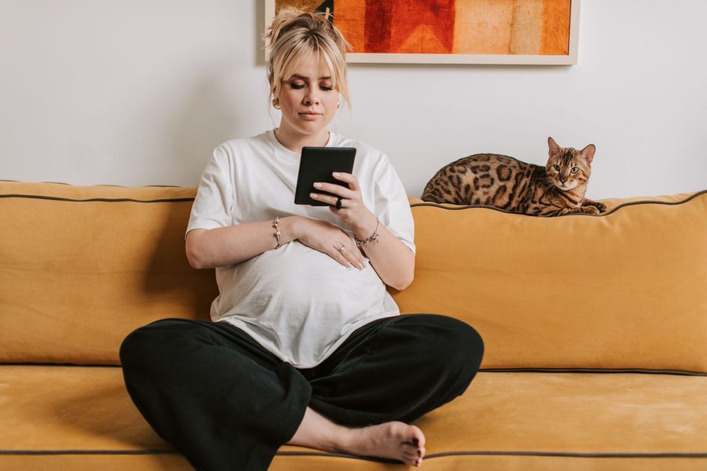 Pregnant woman relaxing on a sofa with a Bengal cat nearby, reading a tablet in a cozy indoor setting.