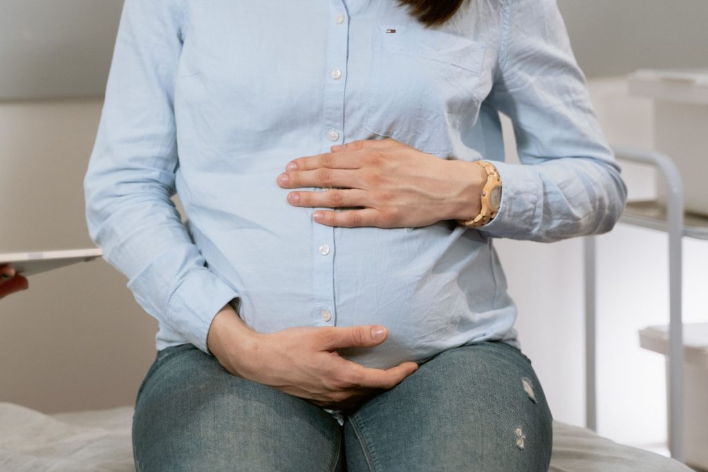 Pregnant woman sitting indoors, hands on belly, wearing casual clothes. Healthcare consultation setting.