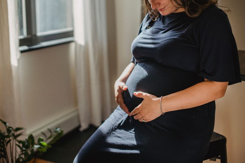 Crop anonymous pregnant woman in blue dress touching belly and looking down while resting in living room in sunny day