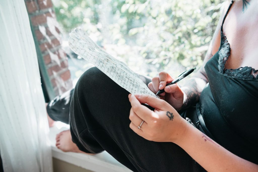 A woman sits on a windowsill writing on paper, bathed in sunlight.