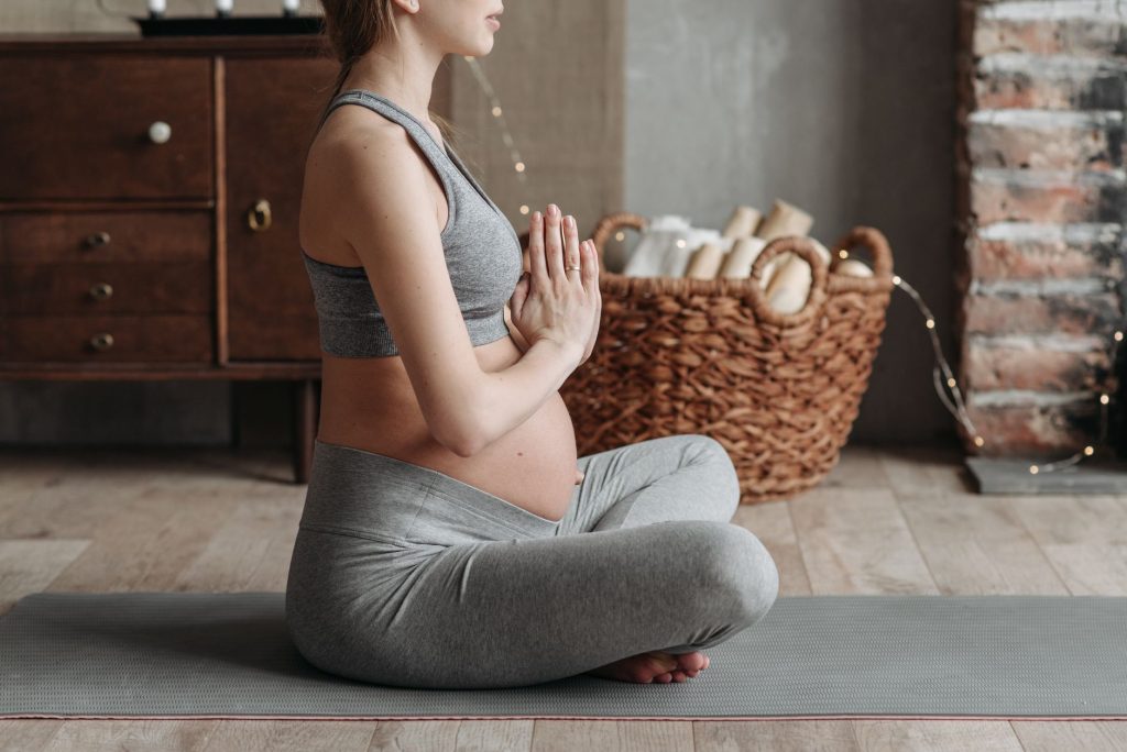 Side view of a pregnant woman practicing meditation on a yoga mat indoors, showcasing calm and wellness.