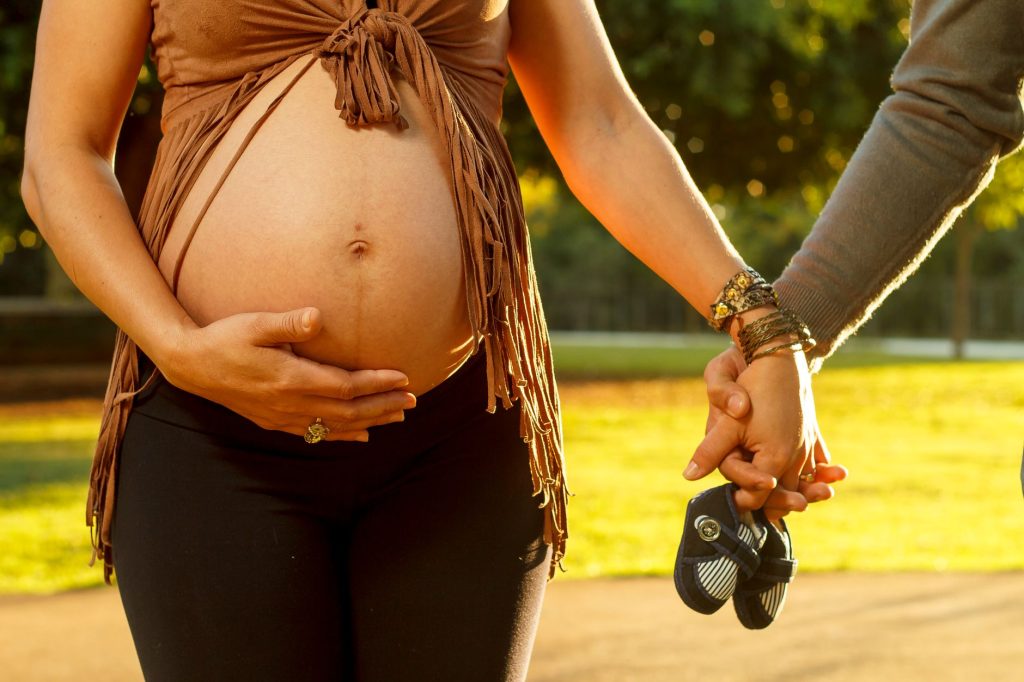 Close-up of a pregnant woman and her partner holding baby shoes in a sunny park.
